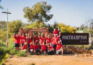 Group of young brokers in red shirts smiling and waving in front of the Northampton town sign, symbolising community support and youth engagement.