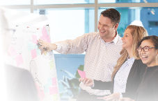 Two people smiling and pointing at sticky notes on a board during a meeting, with a bright, modern office background. Another person smiling holds a pink note.