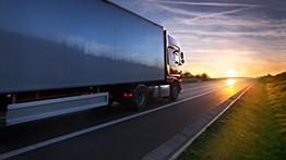 A large truck drives down a highway at sunset with grass on the side.