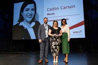 Three people on stage smiling; woman in center holding award. Screen behind shows &#39;Caitlin Carson, Marsh, Congratulations&#39; with Vero logo.