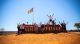 Group celebrating in front of Welcome to Alice Springs sign under blue sky.