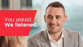 Person in suit smiling with red background text &#34;You asked. We listened.&#34; and blurred cityscape in window behind.